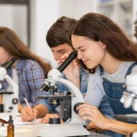 Three students look into microscopes in a science lab classroom