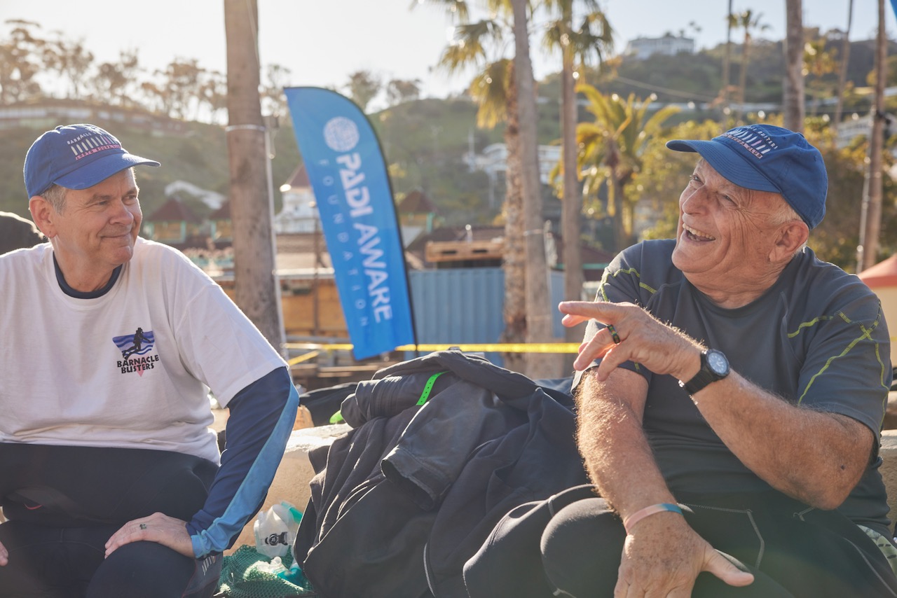two men laugh while sitting in front of a PADI AWARE flag