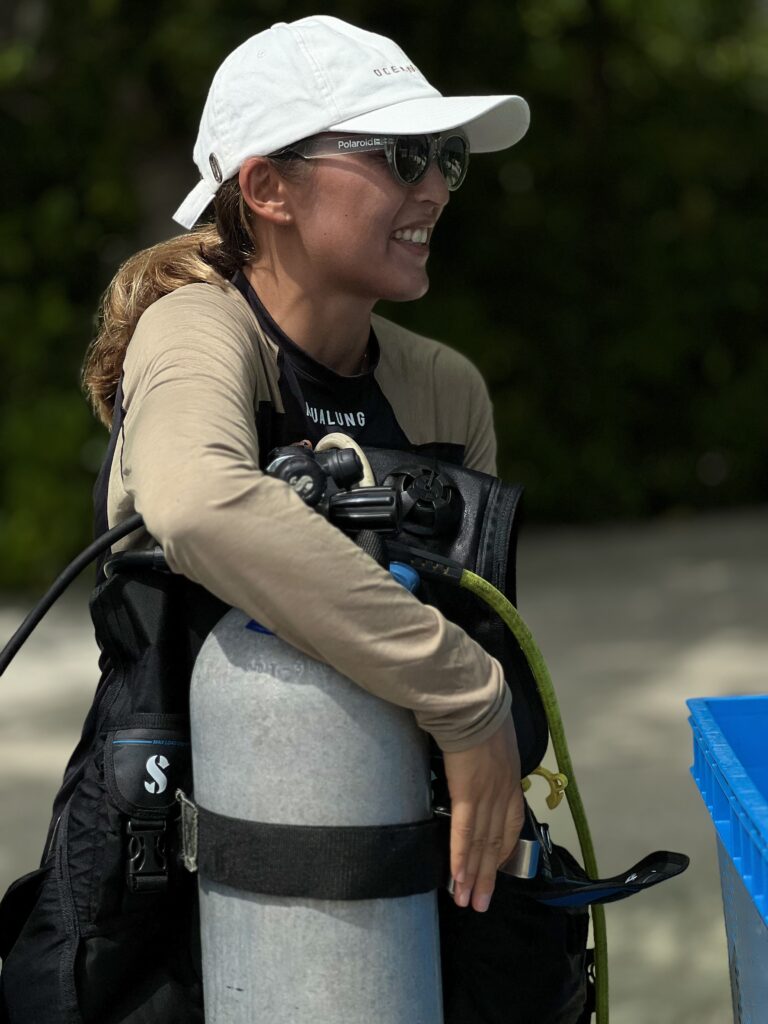 Assyl Yerbolatova smiles as she leans on a tank and scuba kit in the Maldives. She wears sunglasses and a white baseball cap.