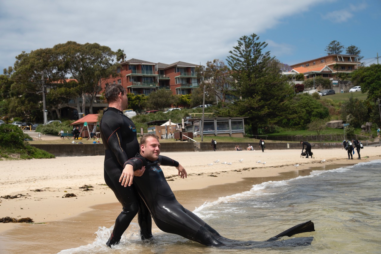 One person in a wetsuit drags another person in a wetsuit out of the water