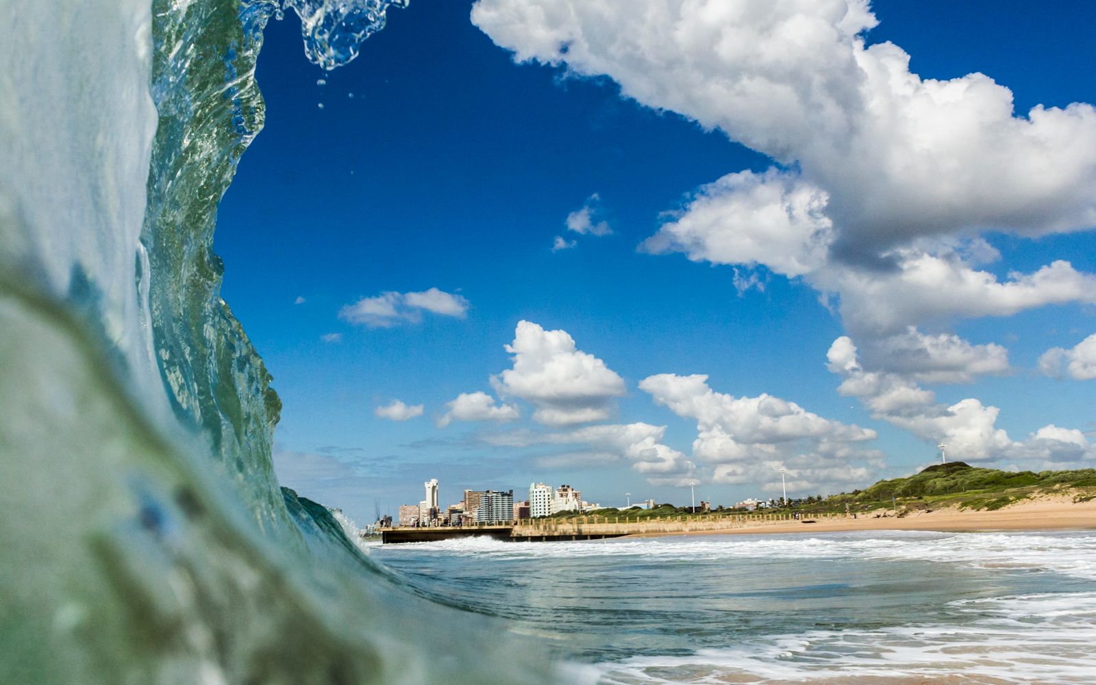 The Durban coastline and white fluffy clouds against a blue sky, as seen through the barrel of a wave crashing on the shore 