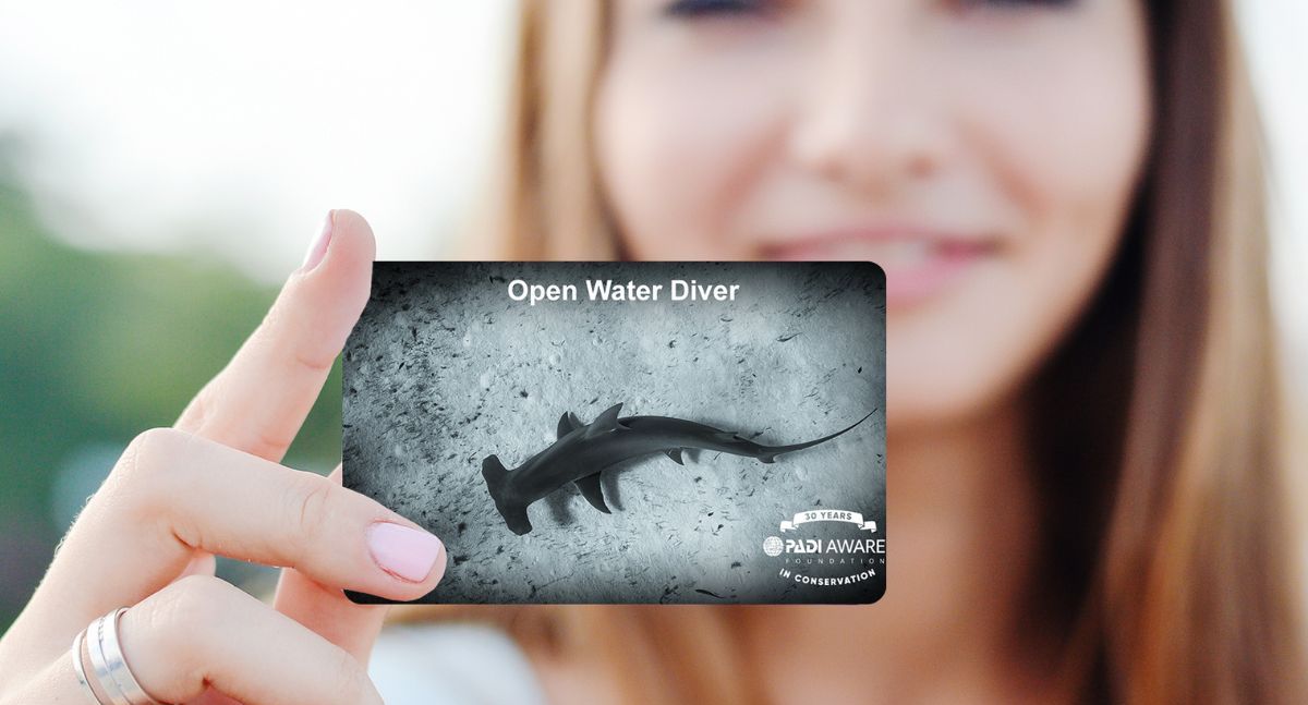 A female diver holds up her PADI AWARE certification card, featuring a hammerhead shark