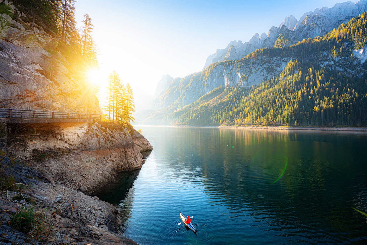 Scenic view of beautiful autumn scenery at famous alpine lake Gosausee with Dachstein mountain summit and young adventurist kayaking in golden morning light at sunrise in fall, Salzkammergut, Austria