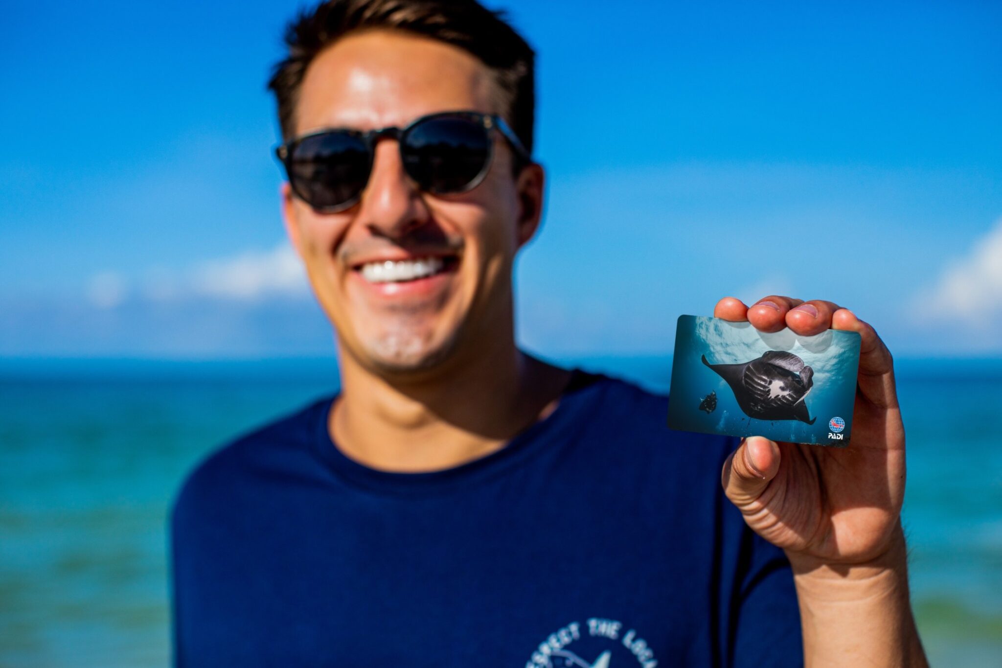 A man on the beach smiles for the camera and holds up a padi certification card showing a manta ray