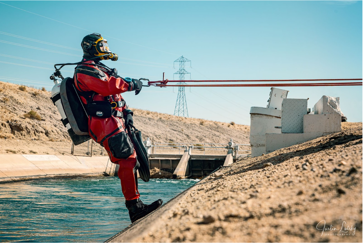 A public safety diver in a protective red suit lowers into a canal for a vehicle extraction mission.