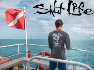 A man wearing a salt life tshirt stands on the side of a boat looking out to sea