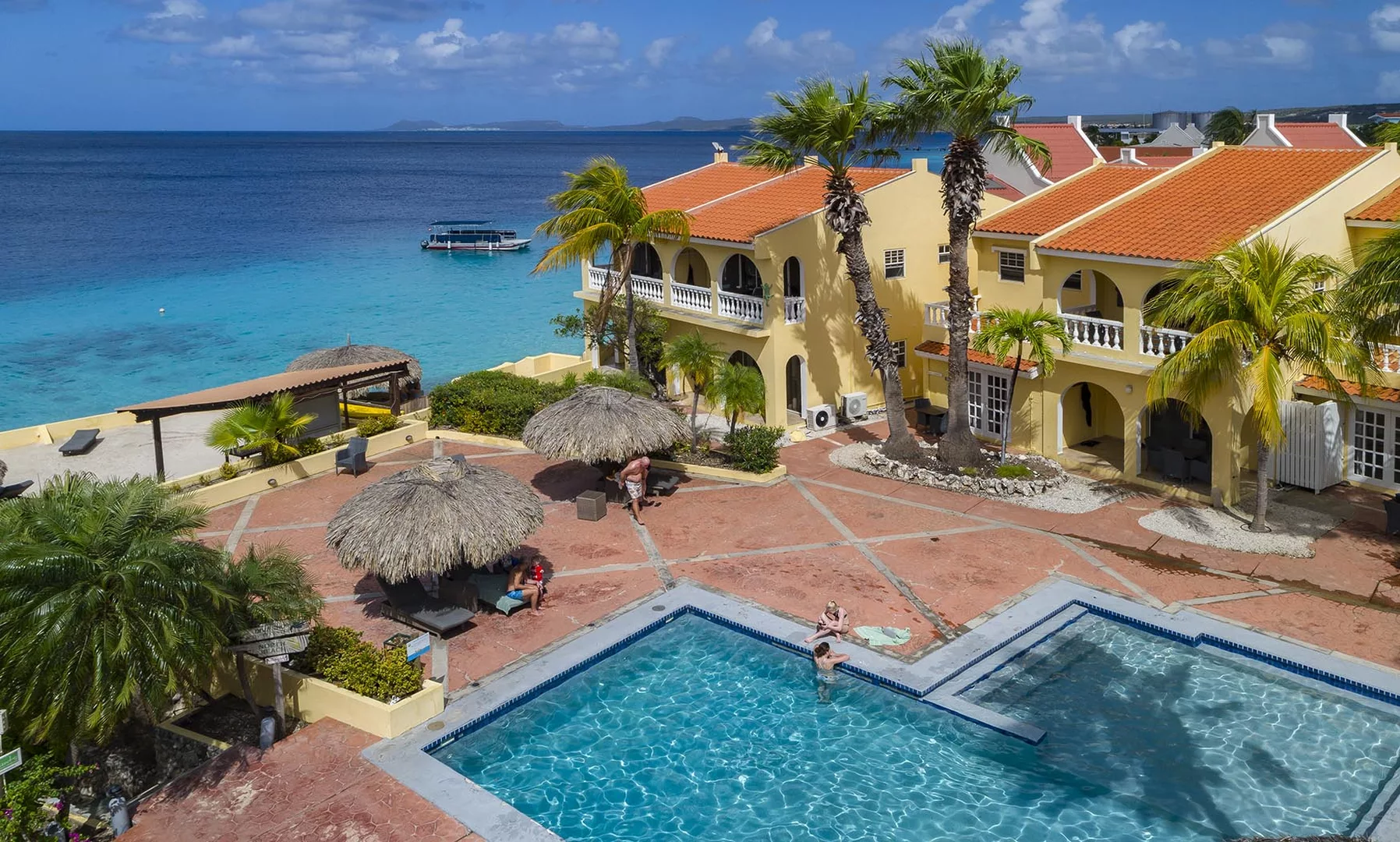 An aerial view of Buddy Dive Resort Bonaire showing yellow painted buildings, a pool, two palapas and the Caribbean Sea