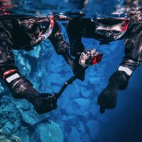 Snorkeling couple young man and woman taking pictures and filming underwater with action camera pointing out to the bottom, blue background crystal clear blue glacial water people wearing dry suits