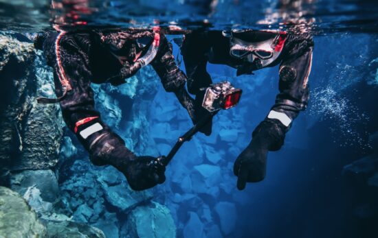 Snorkeling couple young man and woman taking pictures and filming underwater with action camera pointing out to the bottom, blue background crystal clear blue glacial water people wearing dry suits