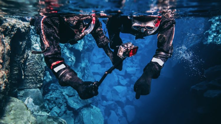 Snorkeling couple young man and woman taking pictures and filming underwater with action camera pointing out to the bottom, blue background crystal clear blue glacial water people wearing dry suits