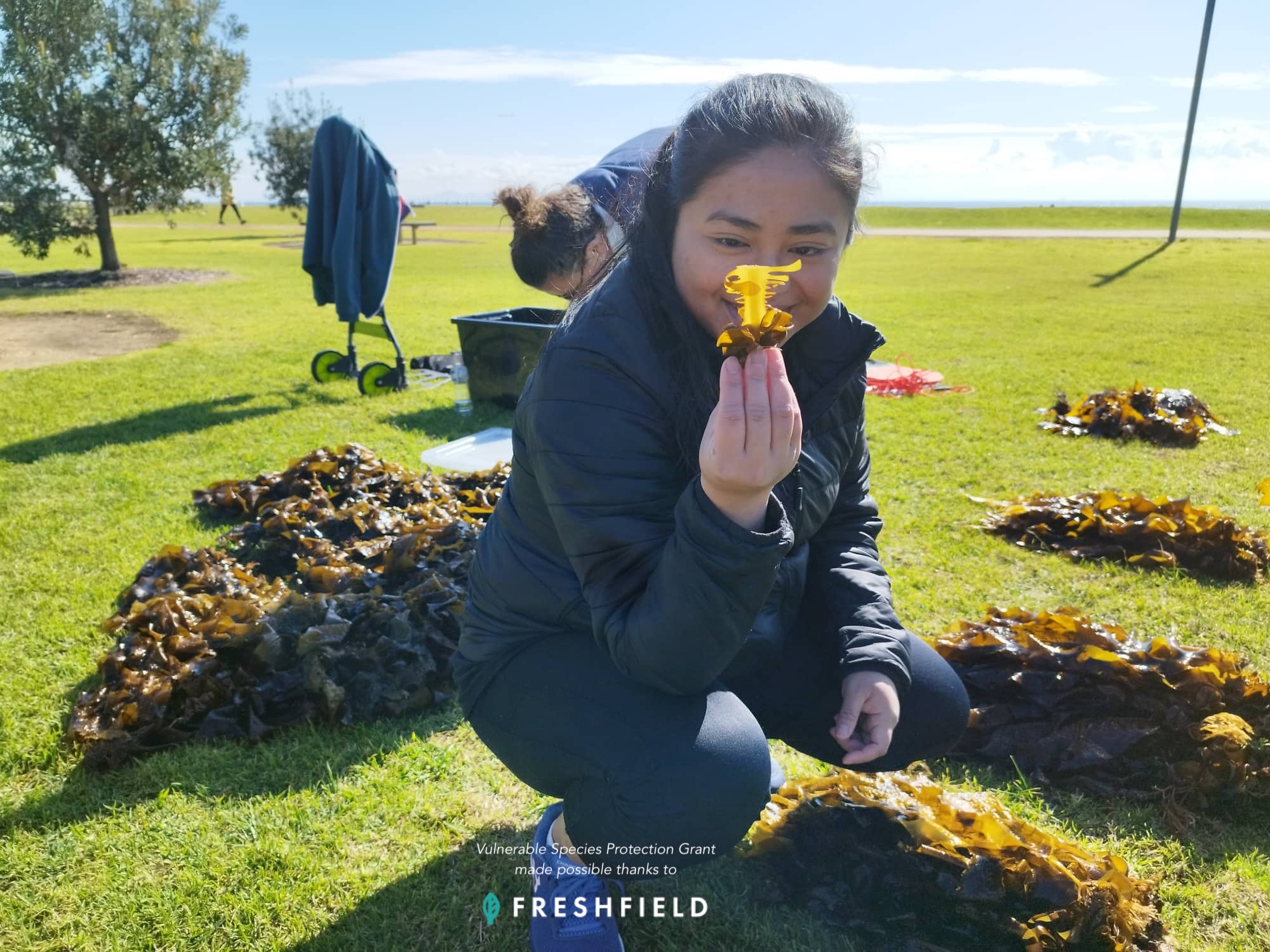 Een vrouw houdt een klein voorwerp omhoog terwijl ze op het gras bij de zee zit