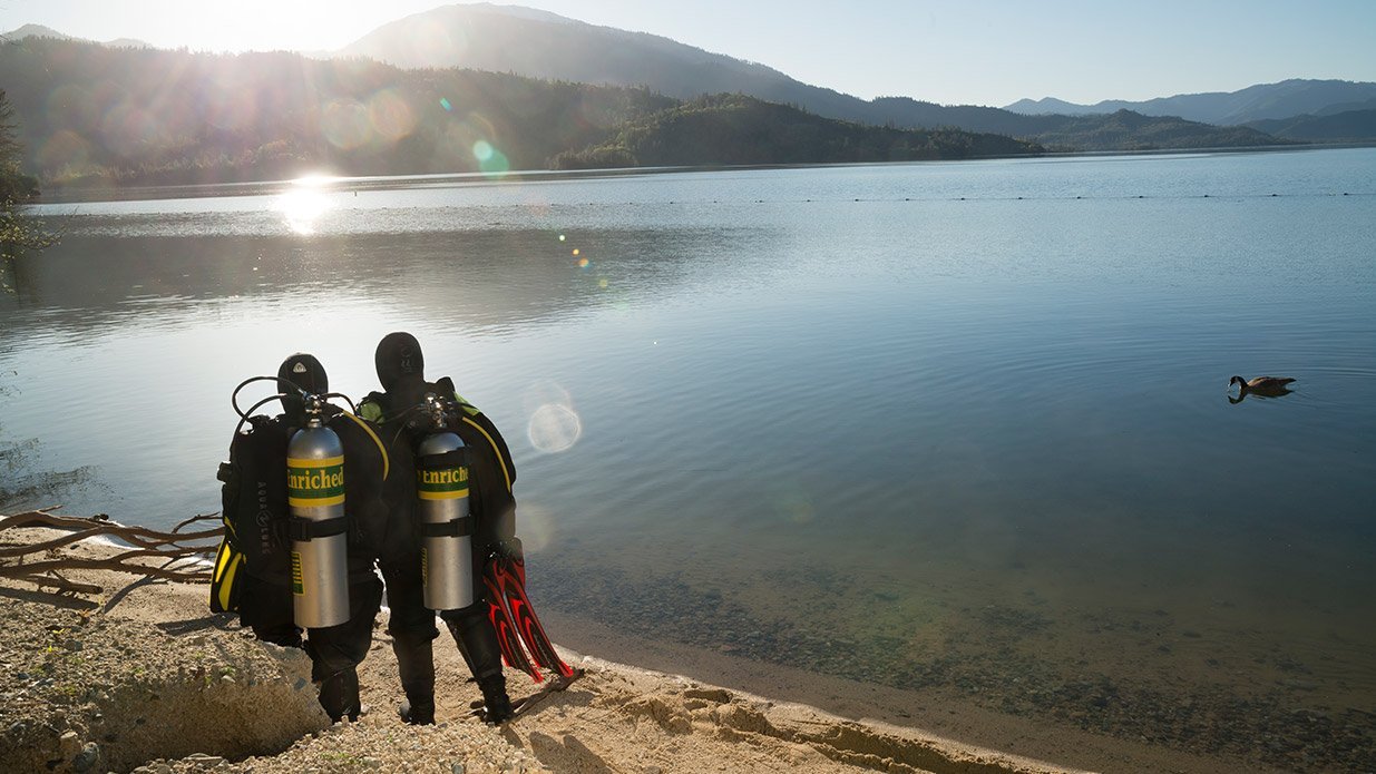 two divers equipped with Nitrox cylinders getting ready to dive into a beautiful lake in a sunny day