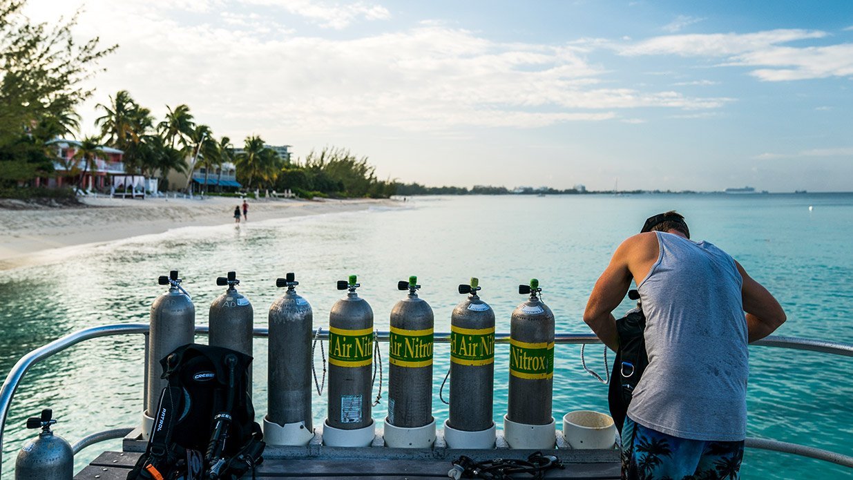 A diver setting up his scuba equipment on a dive boat with enriched air Nitrox cylinders with a beach view on the background