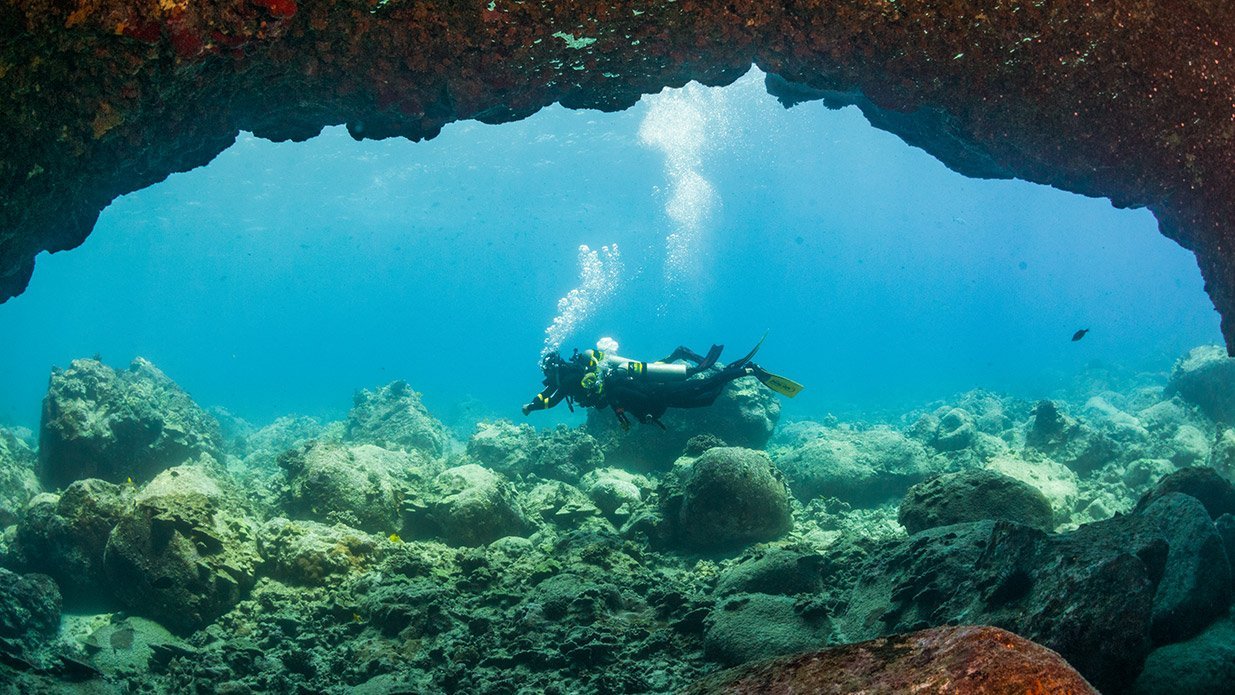 two divers swimming underwater above the rocky bottom using nitrox cylinders