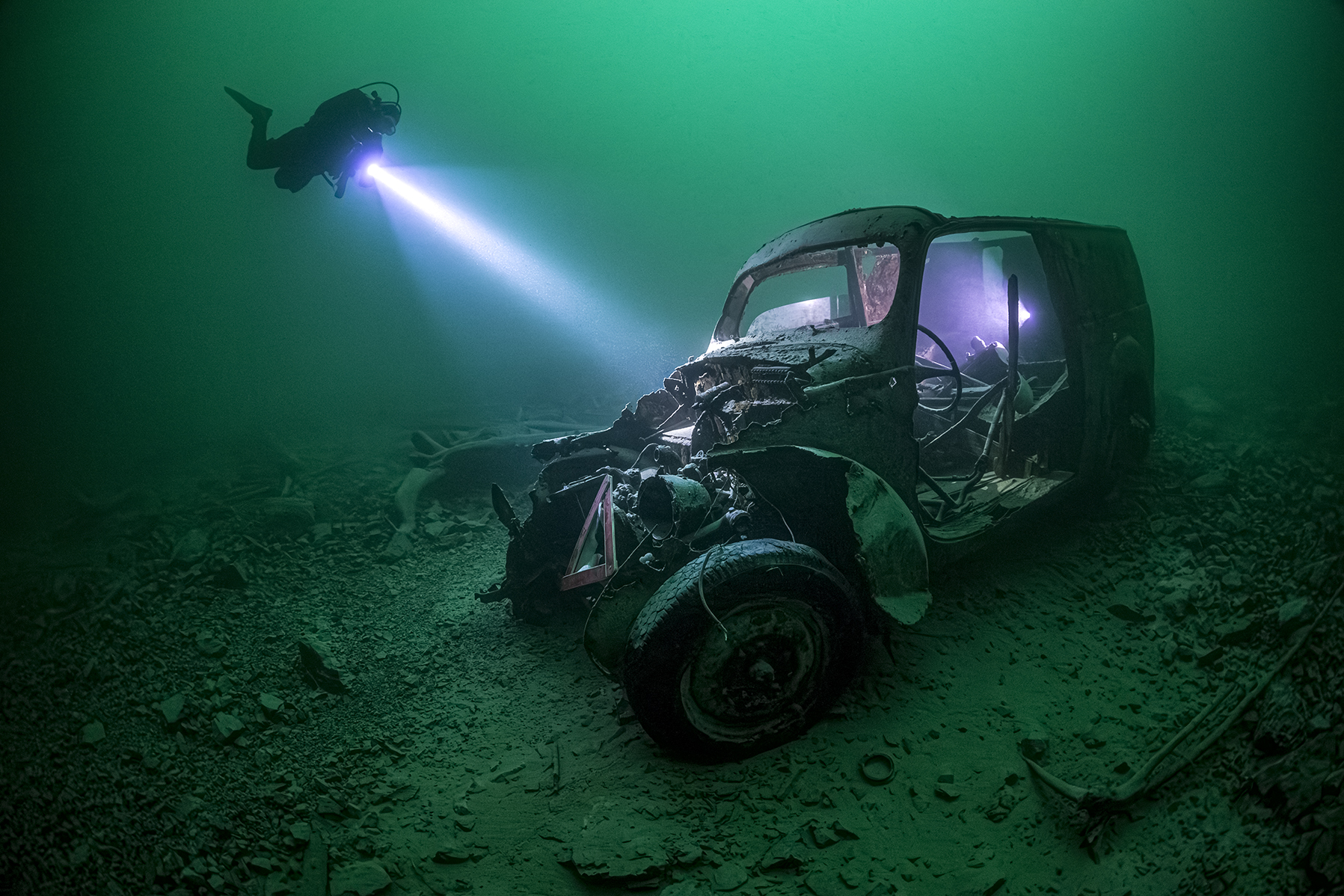 Wreck of a Ford oldtimer from the 50th with a diver holding a dive light towards the car wreck and a light inside the boot, site Galerie, Walchensee, Alps, Germany, PADI.