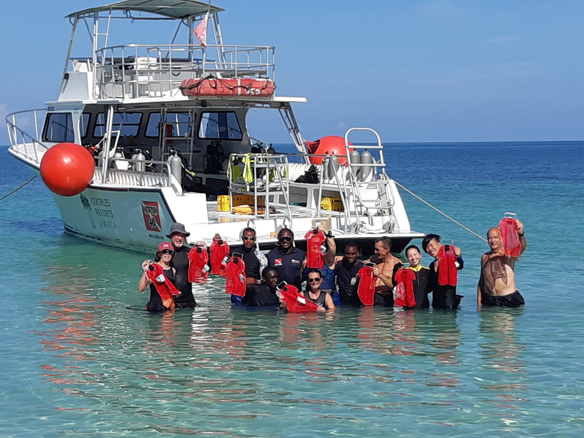 A group of divers stand in shallow water holding up red mesh bags. Behind them is a dive boat.