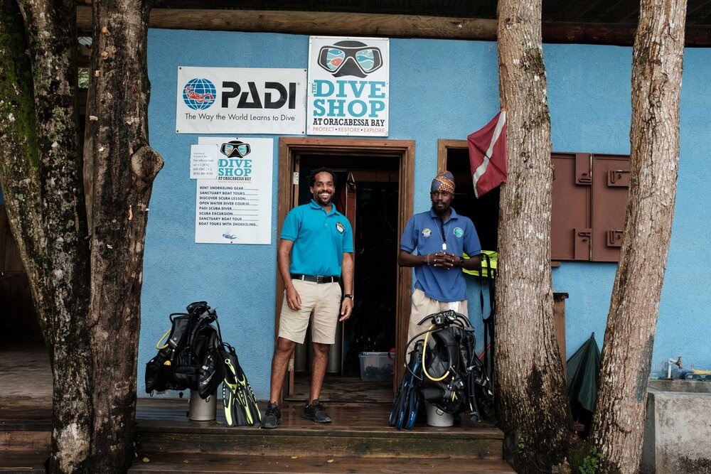 Two scuba instructors stand in front of a doorway with scuba gear at their feet