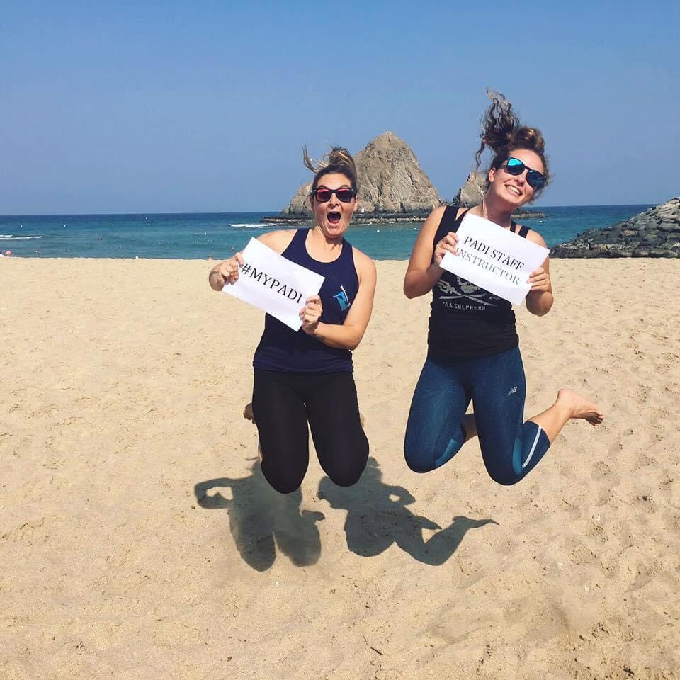 Twee mensen springen en glimlachen op een strand terwijl ze kaarten vasthouden