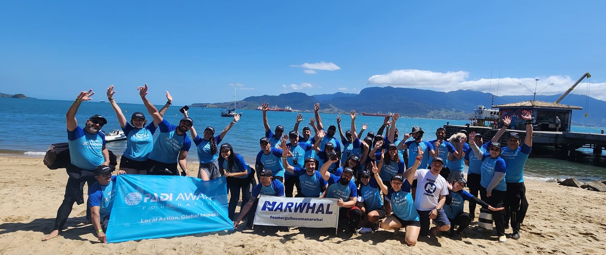 A group photo on a beach in Brazil. About two dozen people wearing turquoise shirts wave at the camera. Two people hold a PADI AWARE banner and others hold a Narwhal Mergulho dive shop banner