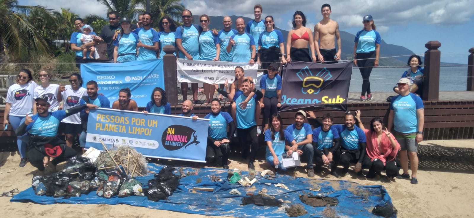 A group photo of smiling people on a beach in Brazil. Several people hold a banner that says "pessoas por um planeta limpo dia mundial dia limpeza." On a tarp in front of them is a pile of debris collected from the ocean