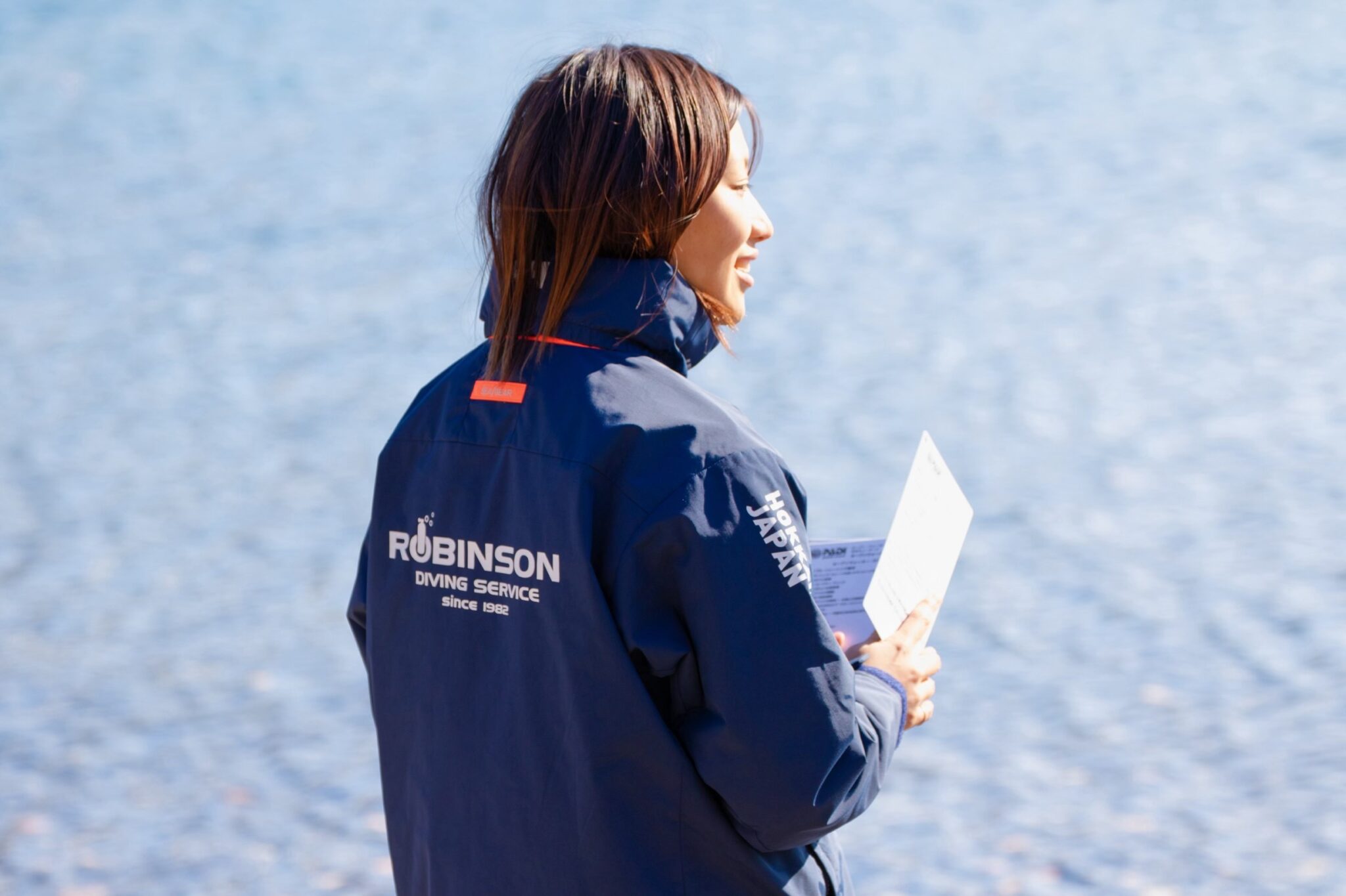 Female instructor wearing a jacket and holding materials in her hand looking out to sea.