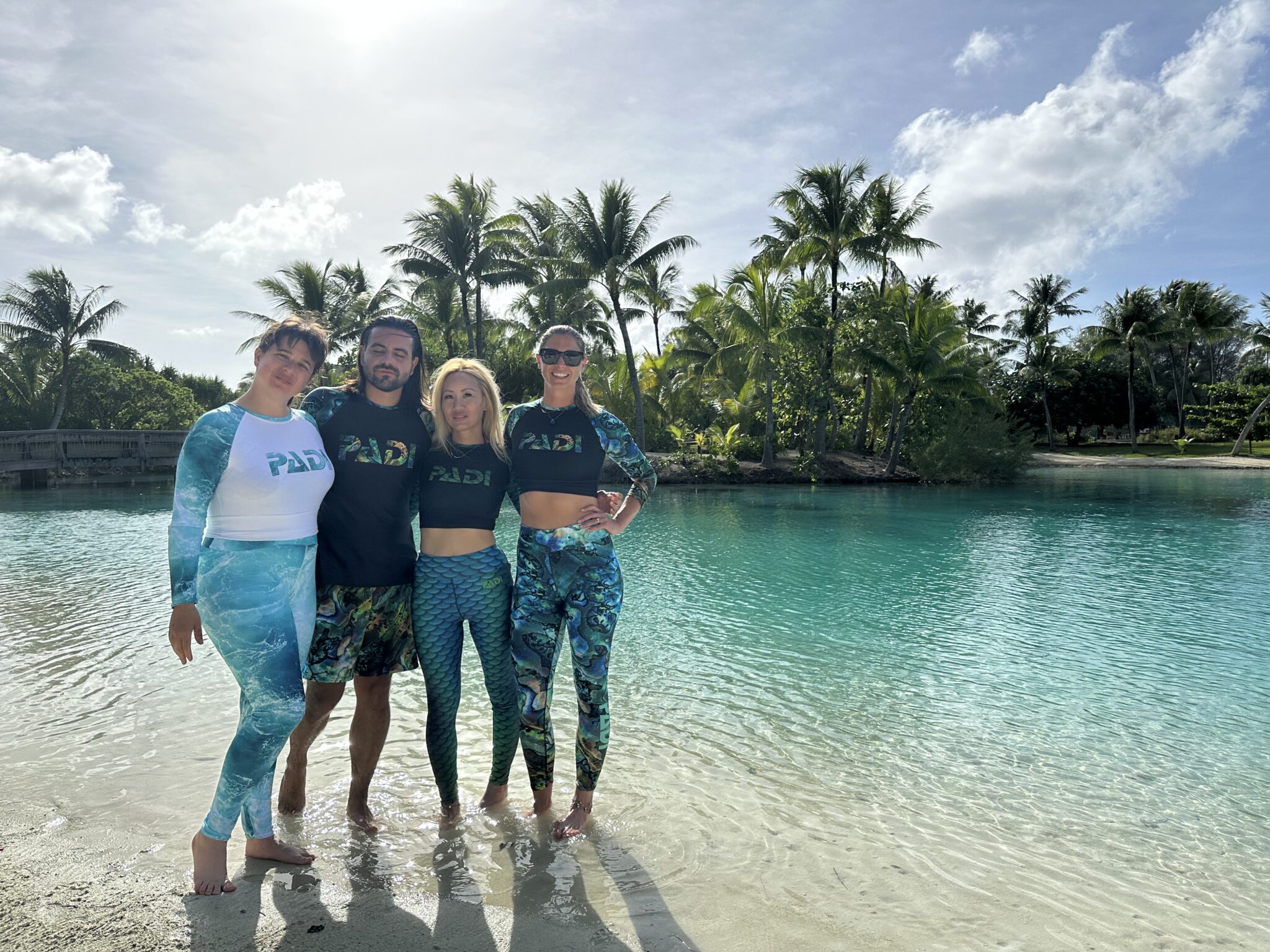 four people stand on a beach in the Maldives. They are wearing PADI gear and smiling for the camera.