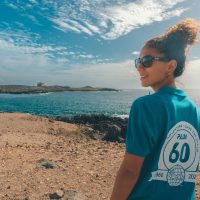 A woman wears a 60th anniversary PADI shirt and walks on a beach, looking back over her shoulder at the camera.