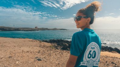 A woman wears a 60th anniversary PADI shirt and walks on a beach, looking back over her shoulder at the camera.