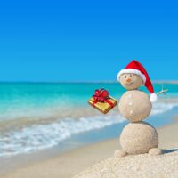 Smiley sandy snowman at beach in christmas hat with golden gift. Holiday concept for New Years Cards.