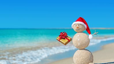 Smiley sandy snowman at beach in christmas hat with golden gift. Holiday concept for New Years Cards.