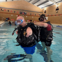 Nic Carapanagiotu donning his scuba gear in a swimming pool with the help of an adaptive support diver