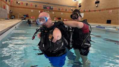 Nic Carapanagiotu donning his scuba gear in a swimming pool with the help of an adaptive support diver