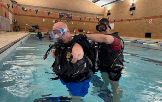 Nic Carapanagiotu donning his scuba gear in a swimming pool with the help of an adaptive support diver