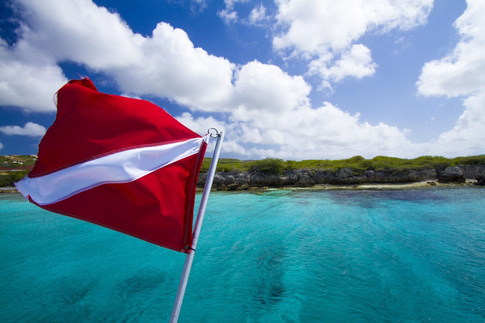 A dive flag flutters above blue Caribbean waters. In the distance, there is a rocky shoreline.