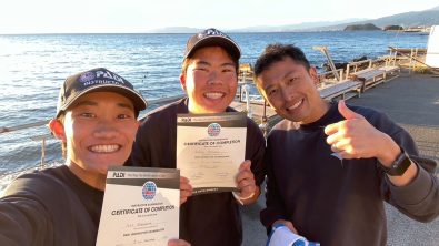 Two newly certified male PADI instructors smile and take selfies with the Examiner with their certificates.