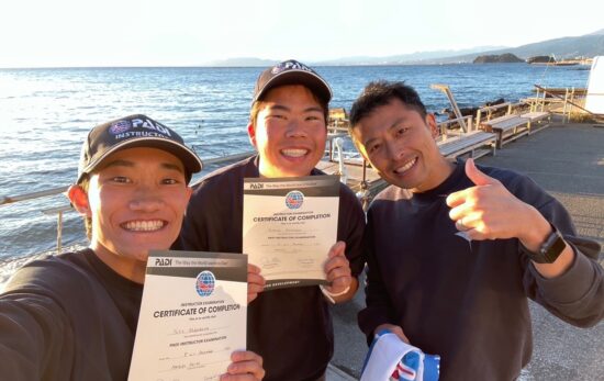 Two newly certified male PADI instructors smile and take selfies with the Examiner with their certificates.