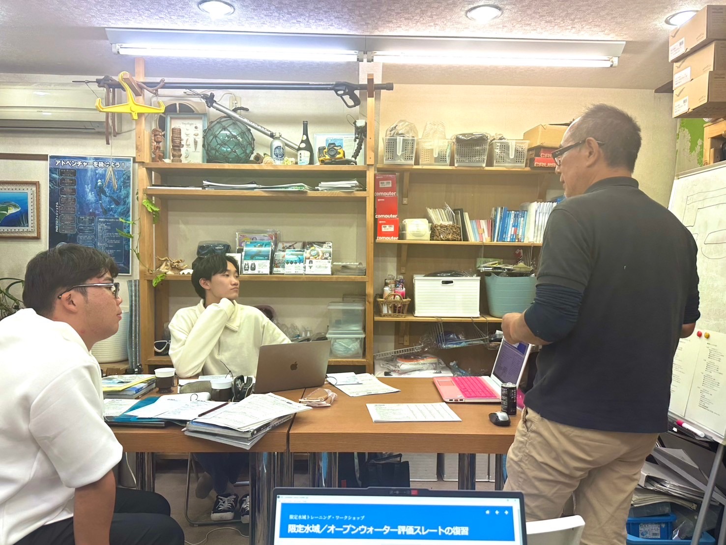 An instructor stands in front of a whiteboard teaching two instructor candidates.