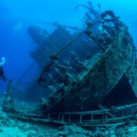 An underwater photographer dives next to an underwater shipwreck lying on it's starboard (right) side.