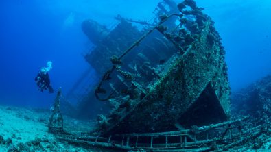 An underwater photographer dives next to an underwater shipwreck lying on it's starboard (right) side.