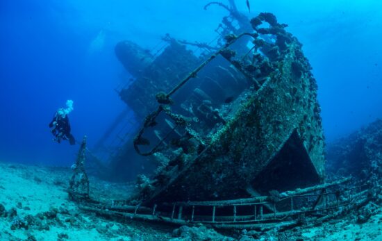 An underwater photographer dives next to an underwater shipwreck lying on it's starboard (right) side.