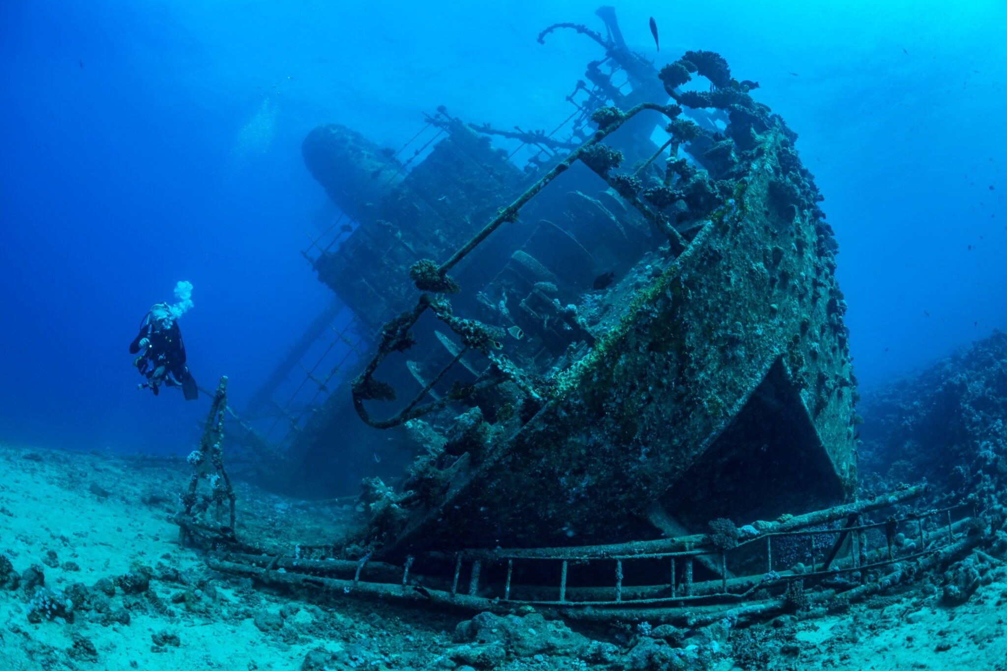An underwater photographer dives next to an underwater shipwreck lying on its starboard (right) side.