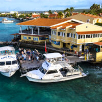 Two dive boats docked at Buddy Dive Resort. Divers stand on the dock waiting to board. In the background is a large yellow building with the dive shop and a restaurant.