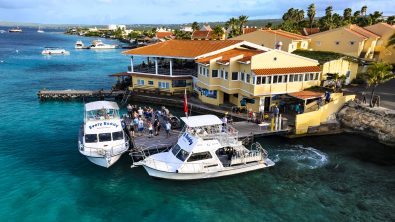 Two dive boats docked at Buddy Dive Resort. Divers stand on the dock waiting to board. In the background is a large yellow building with the dive shop and a restaurant.