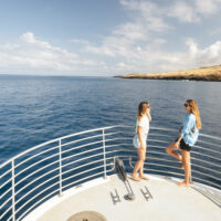 two women stand on the bow of a dive boat. They are talking to each other while wearing Costa sunglasses.