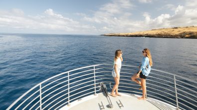 two women stand on the bow of a dive boat. They are talking to each other while wearing Costa sunglasses.