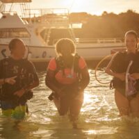 three scuba divers walk out of the water at sunset