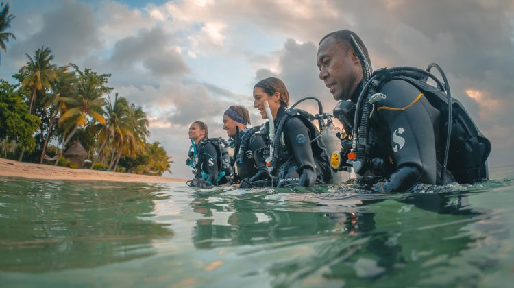 Four divers walking into the water from a beach in Fiji