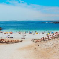 Playa del Virgen beach in Chile. A sandy, fence-lined path slopes down to blue water. A few dozen people relax on the shoreline