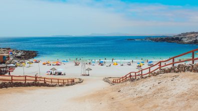 Playa del Virgen beach in Chile. A sandy, fence-lined path slopes down to blue water. A few dozen people relax on the shoreline