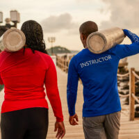 two divers walk down a pier holding tanks on their shoulders